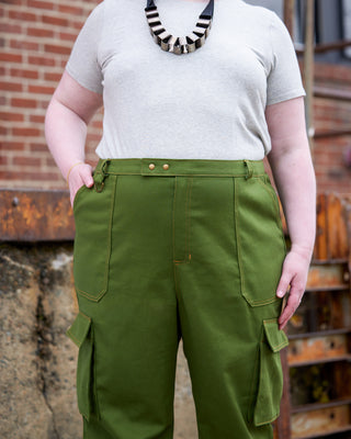 Person wearing green cargo pants and a white shirt with a black and white striped necklace, standing against a brick wall. These pants are the Esmond Cargo Pants sewing pattern from Cashmerette.
