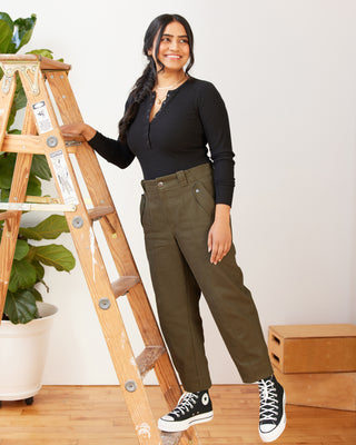 Woman in black top and green pants standing next to a wooden ladder indoors. These pants are the Vinton Pants sewing pattern from Cashmerette.