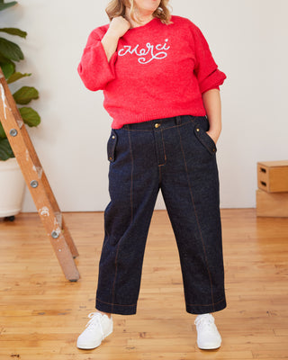 Person wearing a red sweater with 'Merci' text and dark jeans indoors. These pants are the Vinton Pants sewing pattern from Cashmerette.