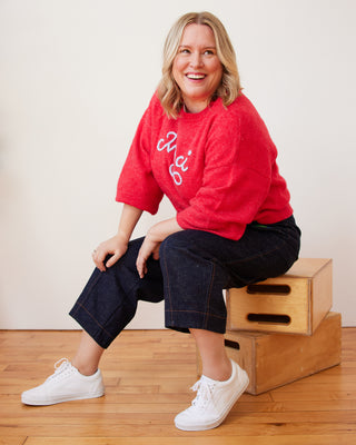 Woman wearing a red sweatshirt with white text, dark pants, and white sneakers sitting on a wooden step. These pants are the Vinton Pants sewing pattern from Cashmerette.