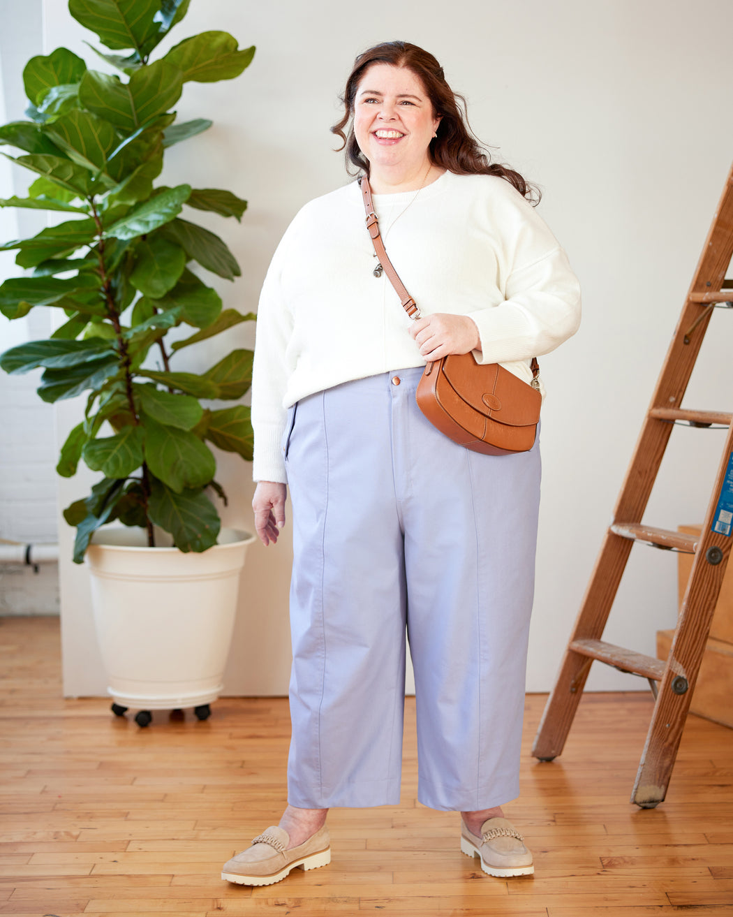 Woman in a white sweater and light blue pants holding a brown leather bag indoors. These pants are the Vinton Pants sewing pattern from Cashmerette.