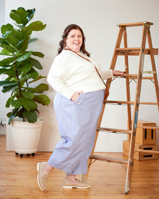 Woman in a white jacket and light blue pants standing next to a wooden ladder in a room with a plant. These pants are the Vinton Pants sewing pattern from Cashmerette.