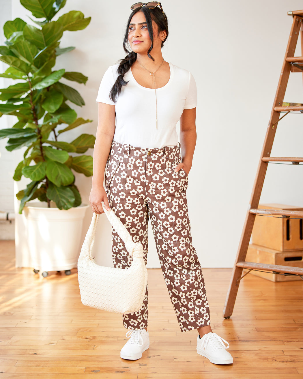 Woman wearing a white top and floral pants standing indoors with a plant and wooden ladder in the background. These pants are the Vinton Pants sewing pattern from Cashmerette.