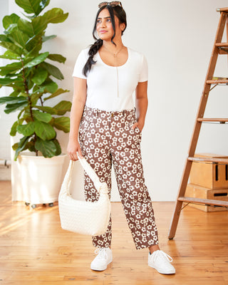 Woman wearing a white top and floral pants standing indoors with a plant and wooden ladder in the background. These pants are the Vinton Pants sewing pattern from Cashmerette.