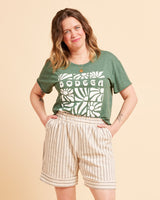 Woman wearing a green t-shirt with 'BODEGA' print and beige shorts on a beige background. These shorts are the Everett Pants & Shorts sewing pattern from Cashmerette.