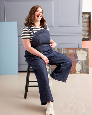 Woman wearing blue denim overalls sitting on a stool indoors. These overalls are the Rhinecliff Overalls sewing pattern from Cashmerette.