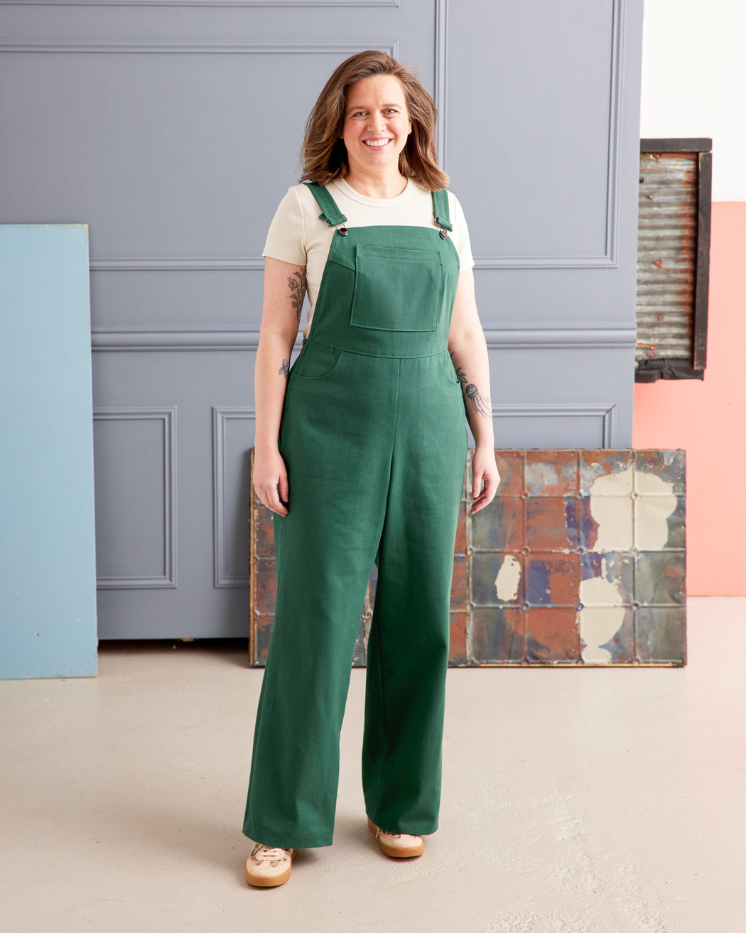 Woman wearing green overalls standing in a room with a gray wall and colorful floor. These overalls are the Rhinecliff Overalls sewing pattern from Cashmerette.