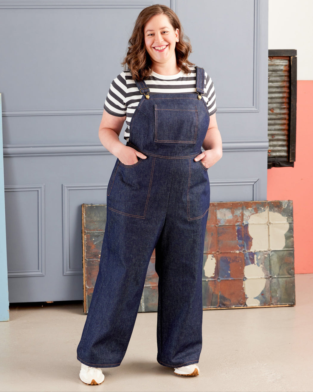 Woman wearing blue denim overalls and a striped shirt standing in a room with a gray wall and colorful abstract painting. These overalls are the Rhinecliff Overalls sewing pattern from Cashmerette.