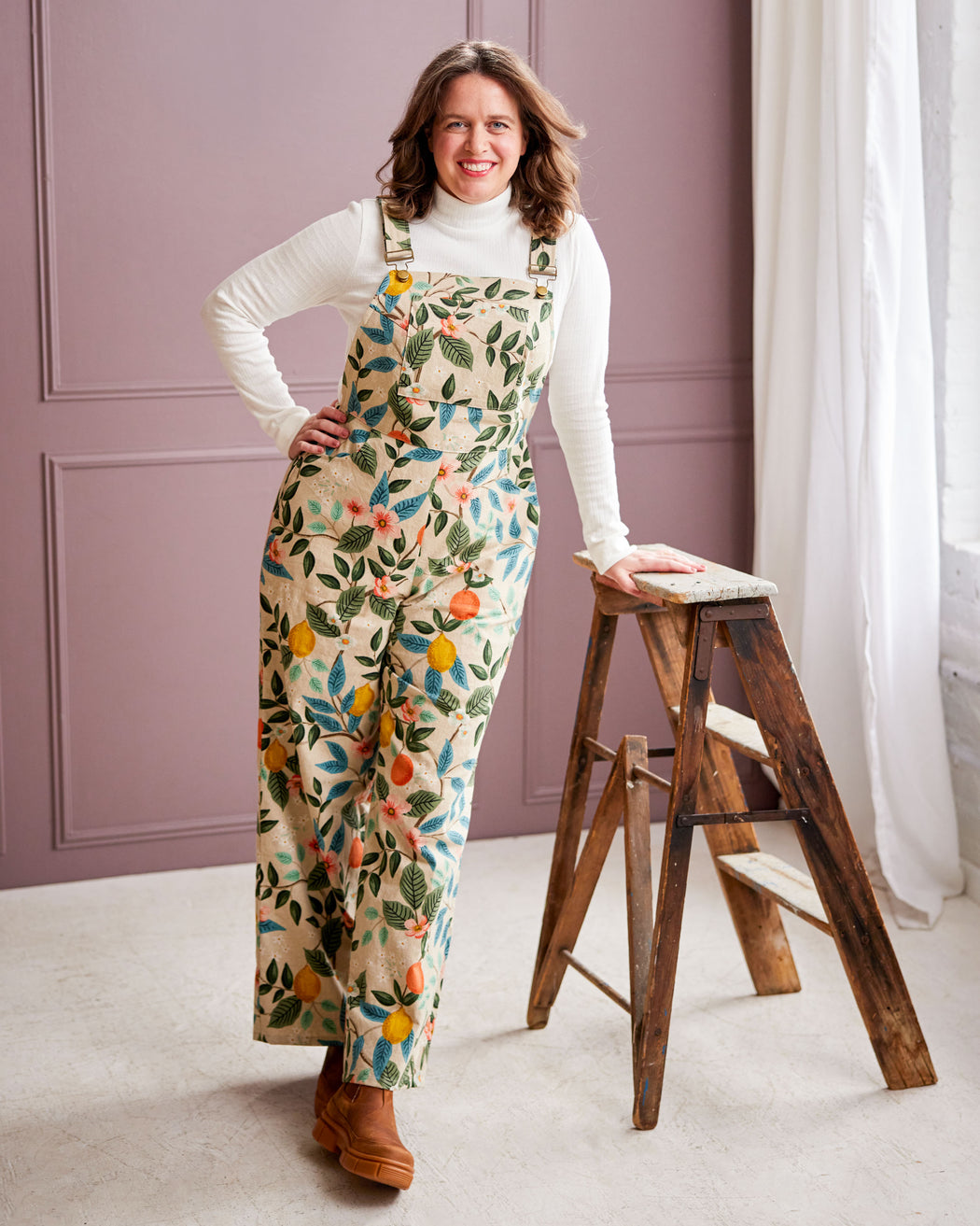 Woman wearing floral overalls standing next to a wooden step stool in a room with a purple wall. These overalls are the Rhinecliff Overalls sewing pattern from Cashmerette.