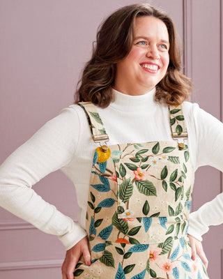 Woman wearing a floral overalls with a white long-sleeve shirt against a purple wall. This is the Rhinecliff Overalls from Cashmerette.