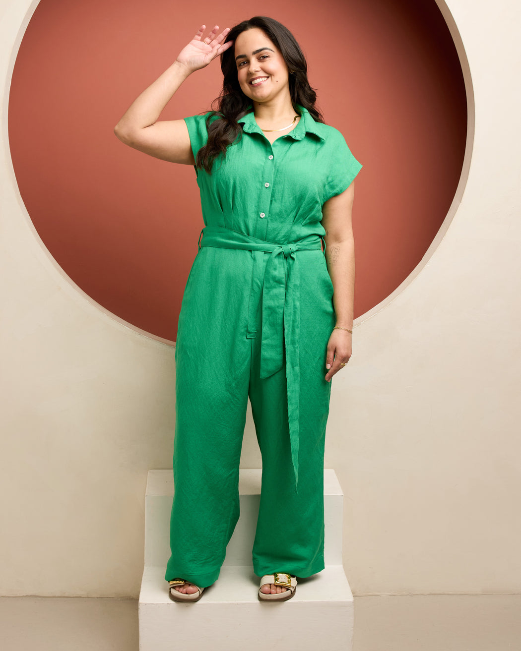 Woman wearing a green jumpsuit standing against a red and beige wall. This is the Roslindale Jumpsuit sewing pattern from Cashmerette.