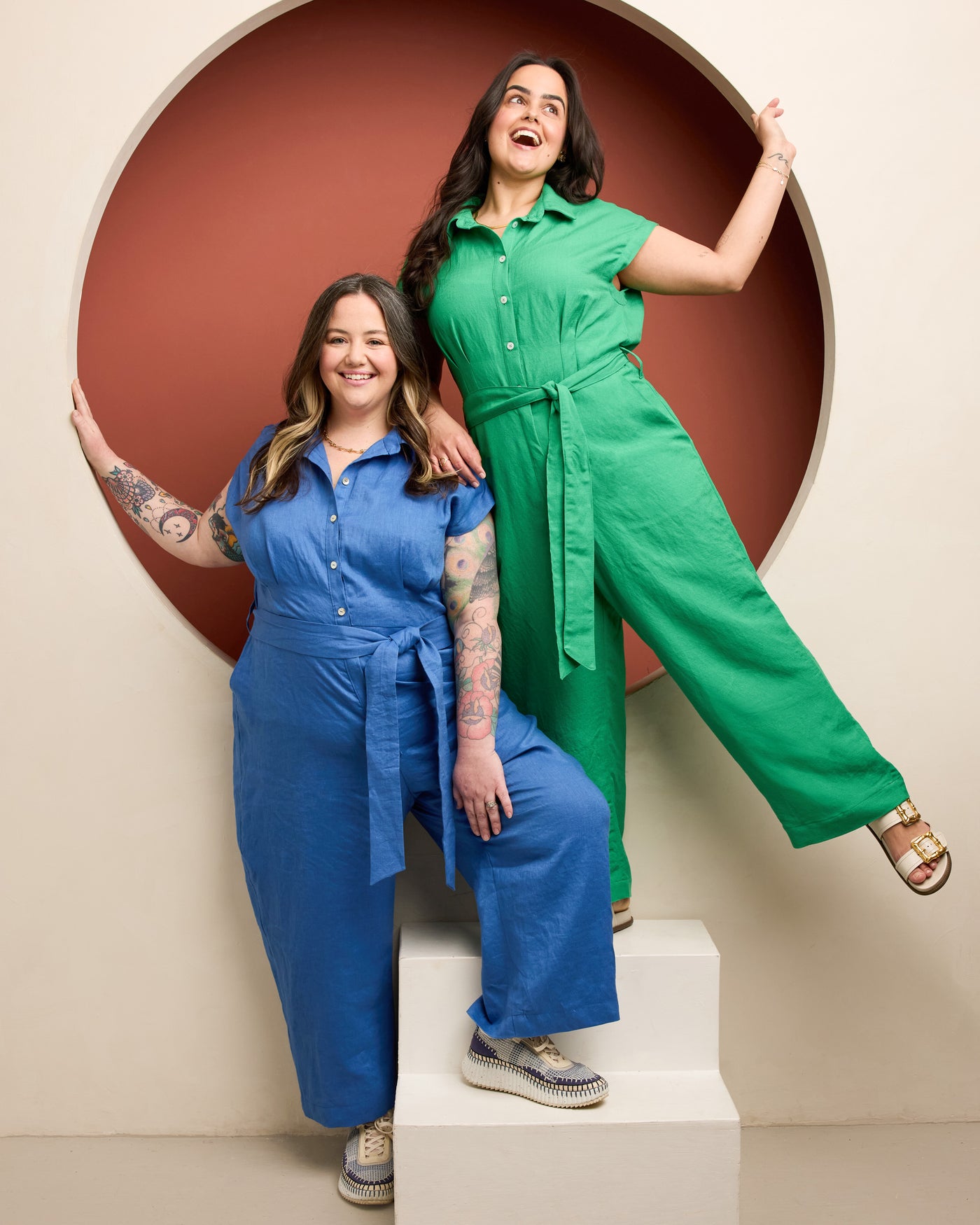 Two women in colorful jumpsuits posing against a beige wall with a circular cutout. This is the Roslindale Jumpsuit sewing pattern from Cashmerette.