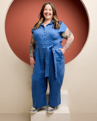 Woman wearing a blue jumpsuit standing on a staircase with a red circular wall in the background. This is the Roslindale Jumpsuit sewing pattern from Cashmerette.