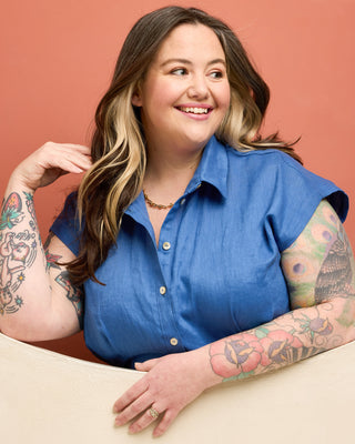 Woman wearing a blue shirt with tattoos on her arms against a peach background. This is the Roslindale Jumpsuit sewing pattern from Cashmerette.