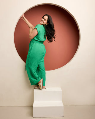 Woman in a green jumpsuit standing on a white platform with a circular red background. This is the Roslindale Jumpsuit sewing pattern from Cashmerette.