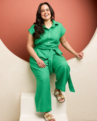 Woman in a green jumpsuit sitting on a white wall against a brown background.  This is the Roslindale Jumpsuit sewing pattern from Cashmerette.