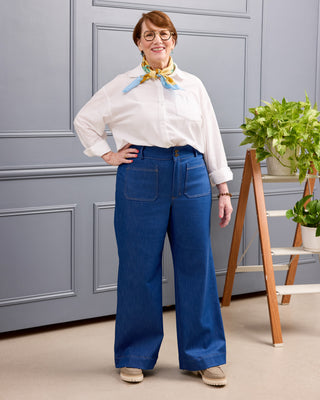 A woman wearing a white long-sleeve button-up shirt, a yellow and blue patterned neck scarf, and high-waisted blue wide-leg jeans, standing in front of a gray paneled wall with a wooden plant stand. This is the Exeter Jeans sewing pattern from Cashmerette.