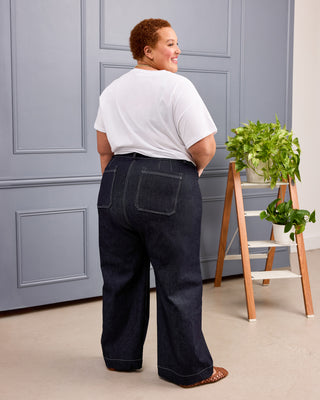 A person shown from the back wearing dark wide-leg denim jeans and a white t-shirt, standing in front of a gray paneled wall next to a small wooden ladder with green plants. This is the Exeter Jeans sewing pattern from Cashmerette.