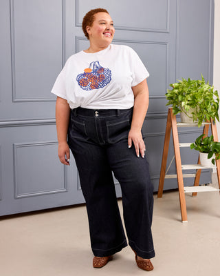 A woman modeling a white graphic t-shirt and dark wide-leg jeans, standing in front of a gray paneled wall with a small plant on a wooden stand. This is the Exeter Jeans sewing pattern from Cashmerette.
