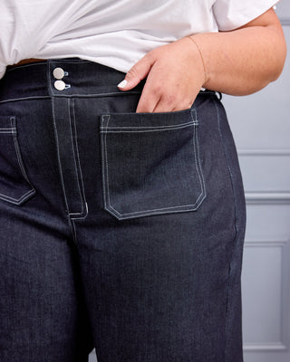Close-up of a person wearing dark indigo denim jeans with white contrast stitching, a white top, and a gold bracelet, standing against a gray paneled wall. This is the Exeter Jeans sewing pattern from Cashmerette.