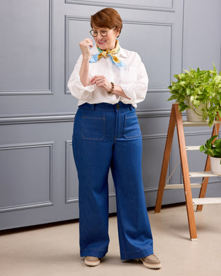 A woman wearing blue wide-leg jeans, a white button-up shirt, and a yellow and blue patterned neck scarf stands in front of a gray paneled wall. She is wearing round glasses and beige shoes, with a wooden ladder shelf holding green potted plants to her right. This is the Exeter Jeans sewing pattern from Cashmerette.
