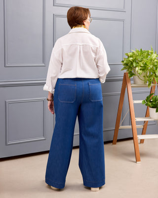 A person viewed from the back wearing a white button-up shirt and wide-leg blue jeans, standing indoors in front of a gray paneled wall with a small wooden plant stand to the right. This is the Exeter Jeans sewing pattern from Cashmerette.