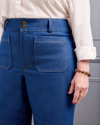 Close-up back view of a person wearing blue denim jeans with a cream blouse, showing the waistband, double button closure, and back pocket details. This is the Exeter Jeans sewing pattern from Cashmerette.