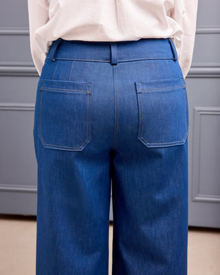 Rear view of a person wearing blue denim wide-leg trousers with a light pink top, standing against a gray paneled wall. This is the Exeter Jeans sewing pattern from Cashmerette.