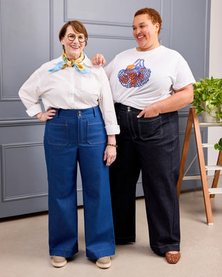 Two women modeling wide-leg jeans in a studio setting with a gray paneled wall background. The woman on the left wears blue jeans with a white blouse and a yellow-and-blue neck scarf. This is the Exeter Jeans sewing pattern from Cashmerette.