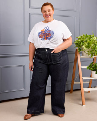 A woman modeling a white graphic t-shirt with a colorful mesh bag print and dark wide-leg denim jeans, standing in front of a gray paneled wall with a wooden ladder shelf holding green plants. This is the Exeter Jeans sewing pattern from Cashmerette.