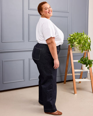 A woman wearing dark blue wide-leg jeans, a white t-shirt, and brown woven shoes stands in profile against a gray paneled wall with a small plant on a wooden stand. This is the Exeter Jeans sewing pattern from Cashmerette.