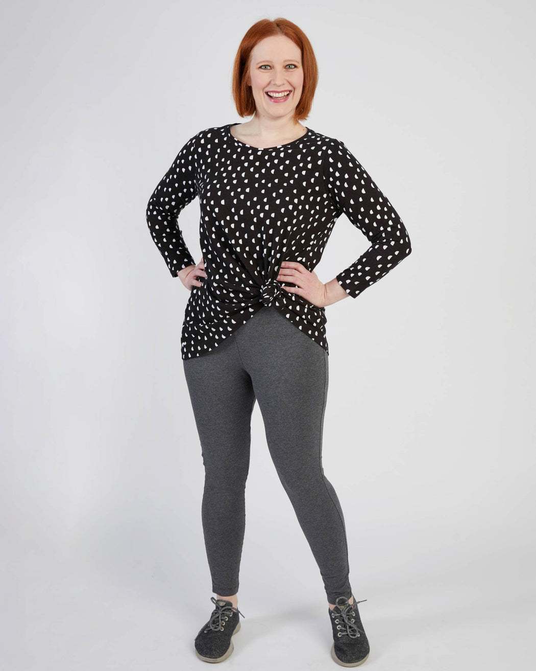 Woman wearing a black polka dot top and gray leggings on a white background. These leggings are the Belmont Leggings and Yoga Pants sewing pattern from Cashmerette.