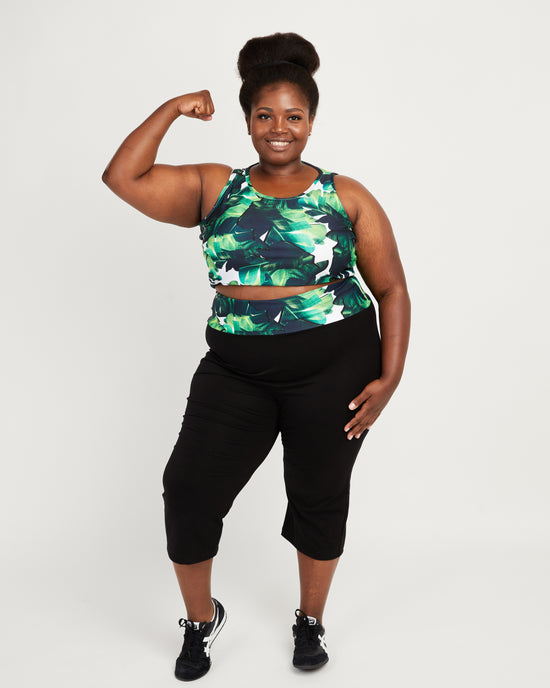 Woman flexing her arm wearing a green and black patterned top and black capri pants on a white background. These leggings are the Belmont Leggings and Yoga Pants sewing pattern from Cashmerette.