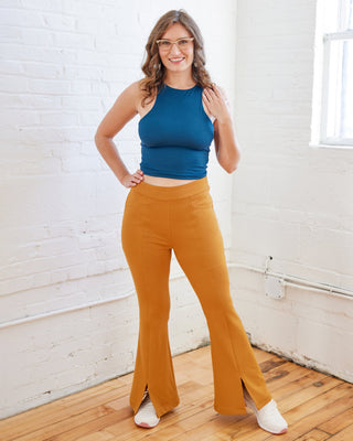 Woman wearing a blue top and mustard yellow flared pants in a room with white walls and wooden floor. These pants are the Ashwood Pants sewing pattern from Cashmerette.