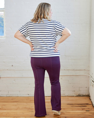 Person wearing a striped shirt and purple pants standing in a room with white brick walls and wooden floors. These pants are the Ashwood Pants sewing pattern from Cashmerette.