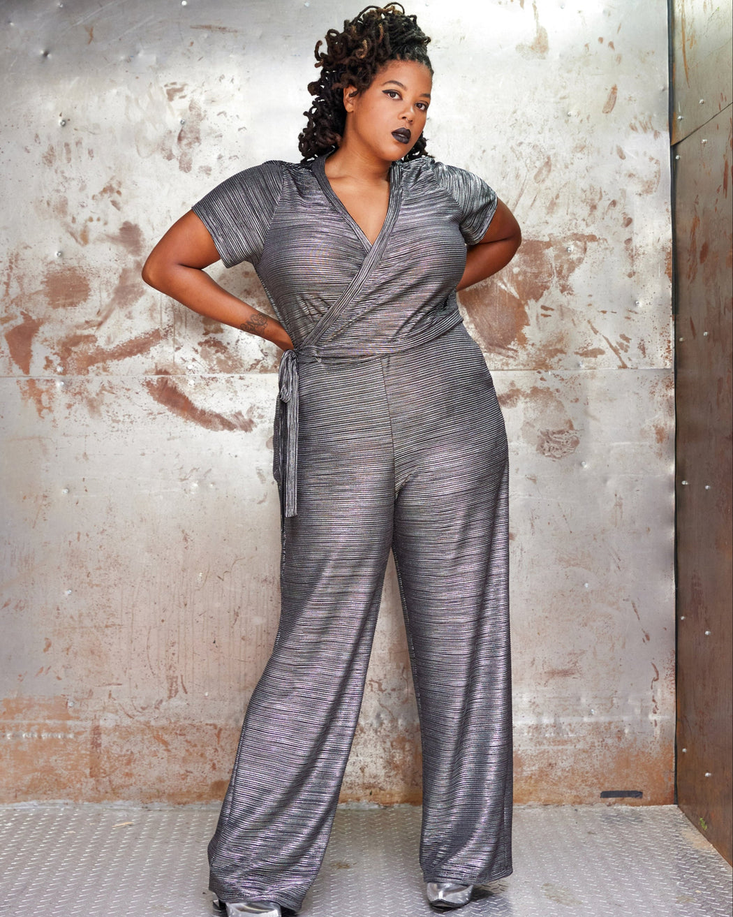 Woman wearing a silver jumpsuit against a textured wall. This jumpsuit is the Mercier Knit Jumpsuit sewing pattern from Cashmerette.