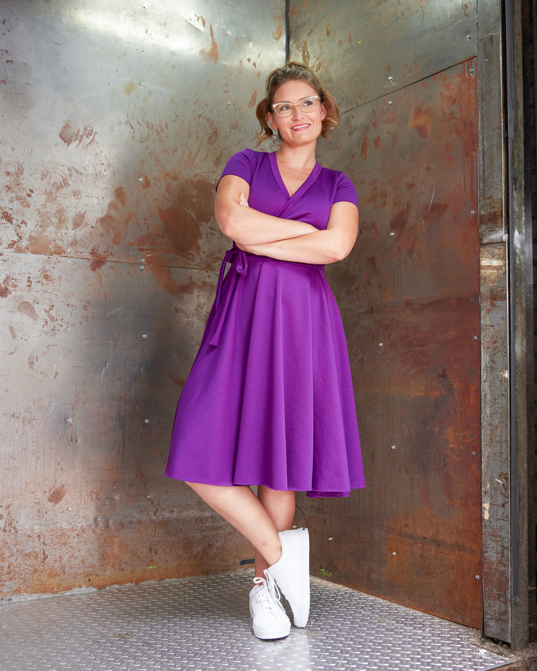 Woman in a purple dress standing against a rusty metal door. This jumpsuit is the Mercier Knit Jumpsuit sewing pattern from Cashmerette.