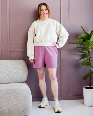Woman in a beige sweater and pink shorts standing in a room with a gray chair and plant. This is the Rowley Shorts sewing pattern from Cashmerette.