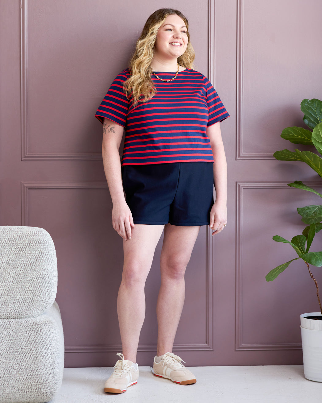 Woman wearing a red and blue striped shirt and navy shorts standing against a purple paneled wall. These are the Wellfleet T-Shirt and Rowley Shorts sewing patterns from Cashmerette.