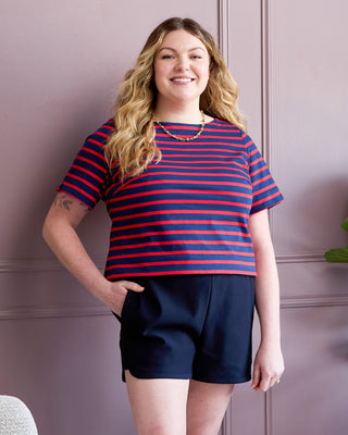 Woman wearing a red and blue striped shirt and navy shorts against a pink wall. This is the Rowley Shorts sewing pattern from Cashmerette.