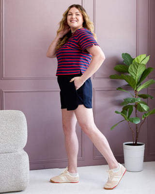 Woman in a striped shirt and shorts standing in a room with a plant and chair. This is the Rowley Shorts sewing pattern from Cashmerette.