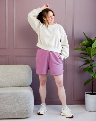 Woman in a white sweatshirt and pink shorts standing in a room with a gray armchair and plant. This is the Rowley Shorts sewing pattern from Cashmerette.