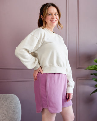Woman wearing a white sweatshirt and pink shorts standing against a purple wall. This is the Rowley Shorts sewing pattern from Cashmerette.