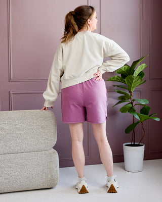 Person wearing a white sweatshirt and pink shorts standing in a room with a gray couch and a plant. This is the Rowley Shorts sewing pattern from Cashmerette.