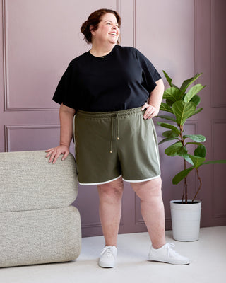Woman wearing a black t-shirt and green shorts standing in a room with a plant and sofa. This is the Rowley Shorts sewing pattern from Cashmerette.