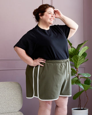 Woman wearing a black t-shirt and green shorts with white trim against a purple wall. This is the Rowley Shorts sewing pattern from Cashmerette.