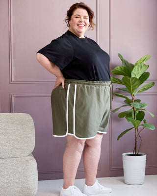 Woman wearing a black top and green shorts standing in a room with a plant and chair. This is the Rowley Shorts sewing pattern from Cashmerette.