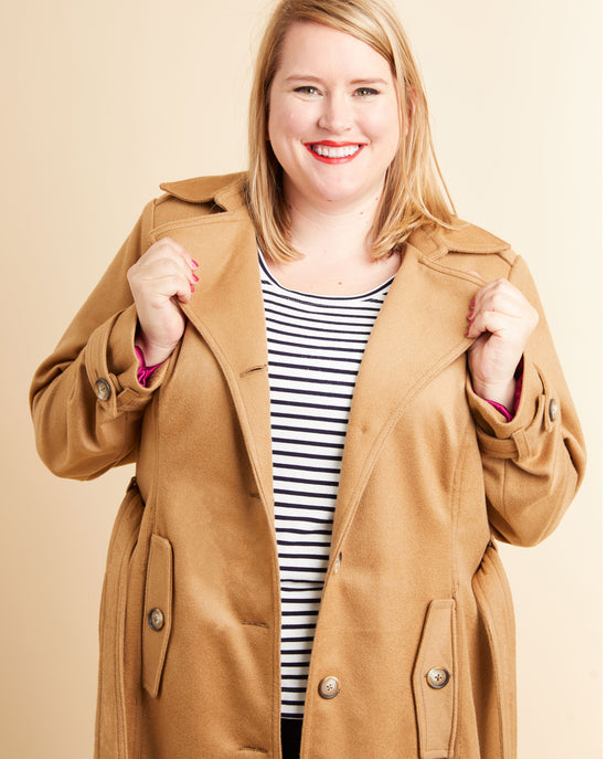 Woman wearing a tan coat over a striped shirt on a beige background. This coat is the Chilton Trench Coat sewing pattern from Cashmerette.