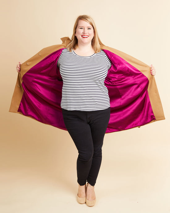 Person wearing a striped shirt and black pants with a magenta and brown jacket on a beige background. This coat is the Chilton Trench Coat sewing pattern from Cashmerette.