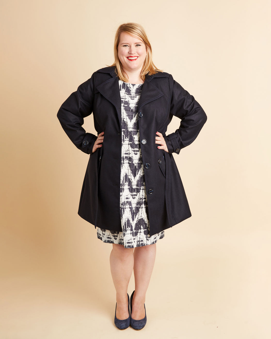 Woman wearing a black coat over a patterned dress on a beige background. This coat is the Chilton Trench Coat sewing pattern from Cashmerette.